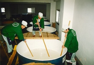 長い棒の先に小さな板をつけた櫂でタンクの中をかき混ぜている3人の杜氏の写