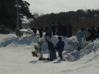雪が積もっている場所に植えた木にむかって、裃を着た男性がおじきしている神事の様子を多くの人たちが見ている写真