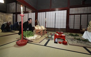 多くのお供え物やお膳が置かれた畳の部屋で紋付き袴の男性が正座しており、後ろの2人が頭を下げている様子の写真