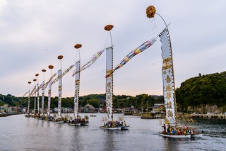 幟と吹き流しを掲げた多くの小型の船が1列に並んで進んでいる写真