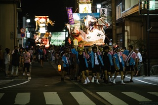 夜に法被を着た人たちが灯りがついたキリコを担いでいる写真