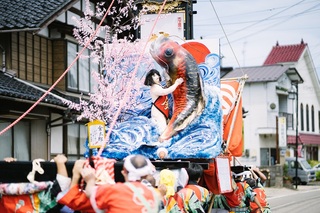 昼間に金太郎と大きな魚が飾られたキリコを法被を着た人たちが担いでいる写真