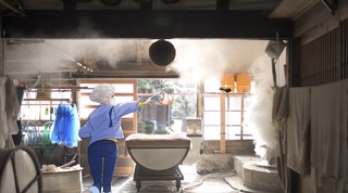 天井に蒸気が立ち込めた作業場に青の作業着を着て、白のフードキャップを被った人のイラストと実際の松波酒造作業場の写真が合成されている画像
