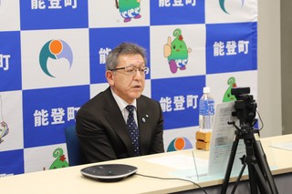 能登町のバックパネルの前で市長が座り、カメラの方向を向いている写真
