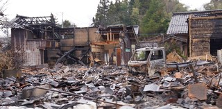 奥には2階建てだったと思われる骨組みの見える住宅と手前には焼けてしまった軽トラック、がれきが散乱している被害の様子を写した写真
