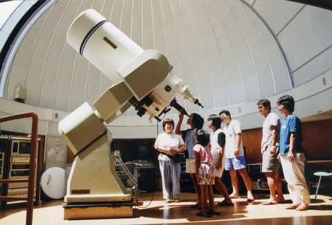 ドーム型の室内の中央に大きな望遠鏡があり、家族が見守る中、一人の子どもが望遠鏡をのぞいている様子の写真