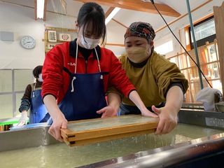 女子生徒が保存会の女性にフォローをもらいながら木枠を持って紙漉き体験を行っている様子の写真