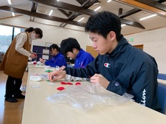 長テーブルに座った3名の生徒が保存会の方に見守られながら作業を行っているコサージュ作りの様子の写真