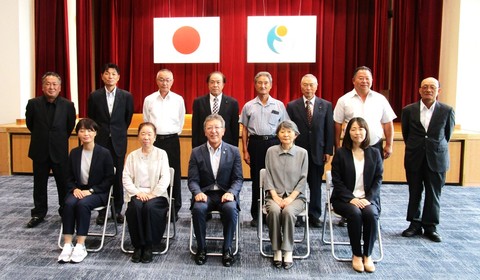 日本国旗と能登町章旗が掲げられているステージ下で前列に椅子に着席した5名と後列に8名が立って記念撮影を行っている写真