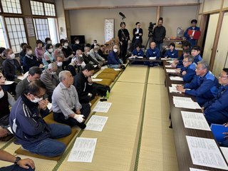 撮影機材が入った会場内で長テーブルをL字型に並べ、その前に青色の作業服を着た関係者複数名が座り、対面になるように大勢の参加者が4列ほどに並んで座り意見交換を行っている様子の写真