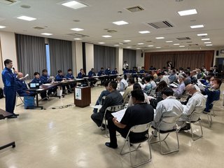 パイプ椅子に座り資料に目を通している大勢の参加者の前で、資料を持ちながら片手にマイクを持って話をしている青色の作業服を着た関係者そして、横並びで長テーブルに座っている関係者十数名が写る意見交換会の写真