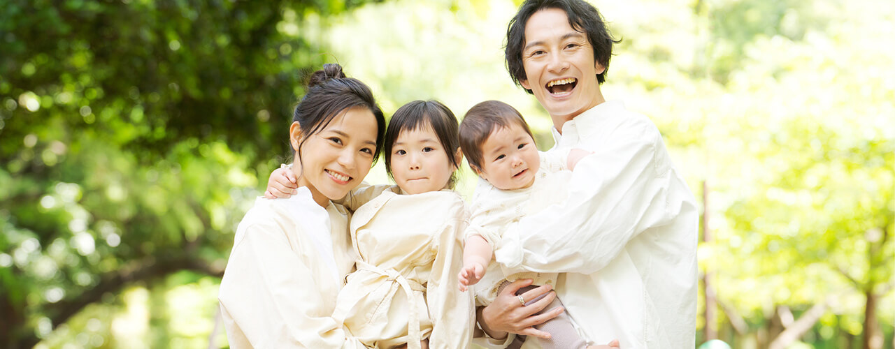 幼い子ども2人を抱きかかえる両親 日が降り注ぐ木々の下で笑っている