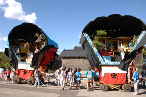 青空の下、人形や竜で飾られた2つの山車の回りに多くの人たちが集まっている写真