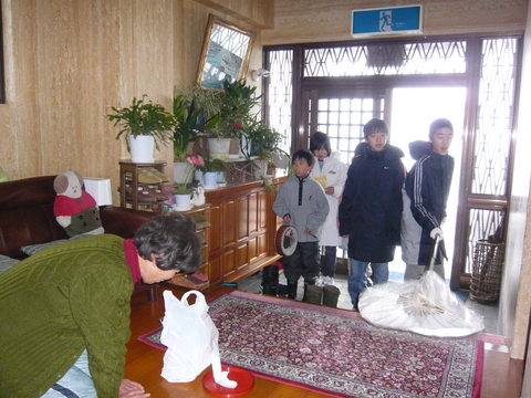 玄関に太鼓や扇をつけた道具を持った子供たちが集まり、家の住人が正座をして頭を下げている写真