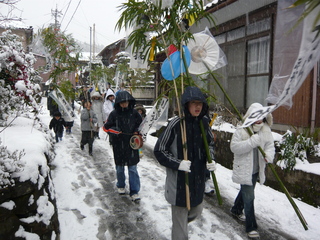 雪道を多くの子供たちが太鼓や扇子のついた竹を持ちながら歩いている写真