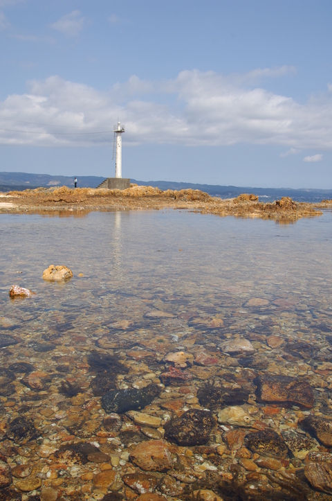 水面から浅瀬の石が見える海沿いから灯台が見えている写真
