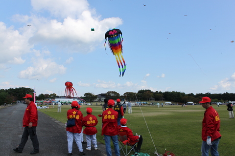 青空の下、芝生がある広場でお揃いの赤の帽子と上着姿の人たちが鮮やかな色の凧をあげている凧あげまつりの様子の写真