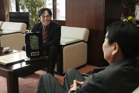表彰盾が机の上に置かれ、高市さんが笑顔で市長に話をしている様子の写真