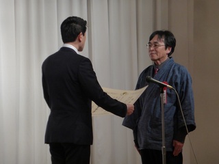 あかま総務大臣政務官が賞状を読み上げ、高市さんが表彰されている様子の写真