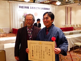 会場の舞台上に「平成26年度ふるさとづくり総務大臣賞」と掲示され、高市さんが賞状を持って前川孝昭名誉教授と記念撮影している写真
