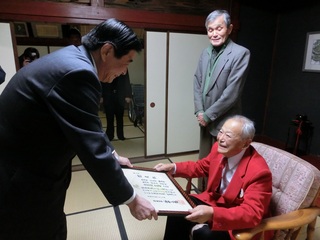 持木町長が渡す認定証を椅子に座って笑顔で受け取る宮本さんの写真