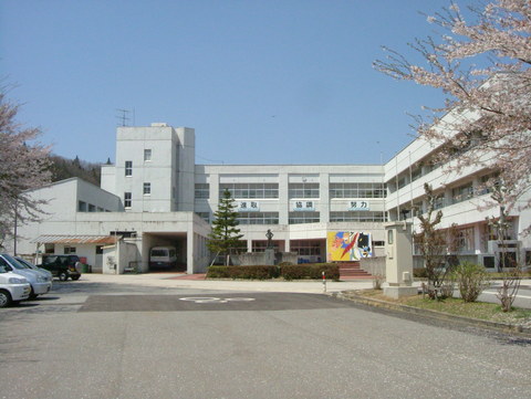 白い外壁で3階建てのL字型の校舎と周囲に桜の木や植栽が施された中学校の外観写真