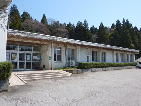 1階建ての白壁で屋根のほうに植物のツタが這っており、入り口前に階段とスロープが設置されている建物の外観写真