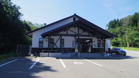 周りは青々とした木々が立ち、三角屋根で白壁の平屋づくりの建物の入り口前に駐車場がある建物の外観写真