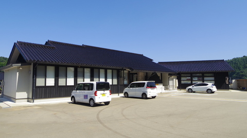 平屋づくりでL字型の建物と建物の前の広々とした駐車場に3台の車が停まっている写真