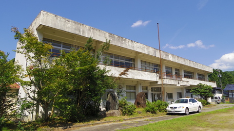 白い外壁で2階建て、建物の前に二つの石碑がありその横に背の高い木が立っている建物の外観写真