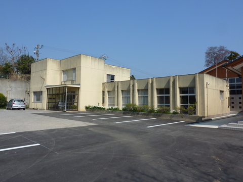 白い外壁で左側が2階建て、右側が大きなガラス窓がある1階建て、建物の前には広々とした駐車場がある外観写真