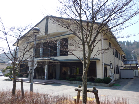 葉が落ちた街路樹と街頭の向こうに建っている、ベージュの外壁で2階建て、三角屋根の公民館の建物の外観写真