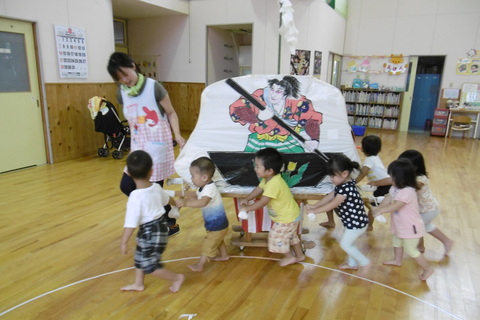 室内で一寸法師の絵が描かれた山車を押している子供たちと先生の写真