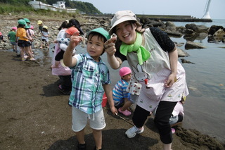 海岸で拾った貝殻を手にもってこちらに見せている男の子と先生の写真