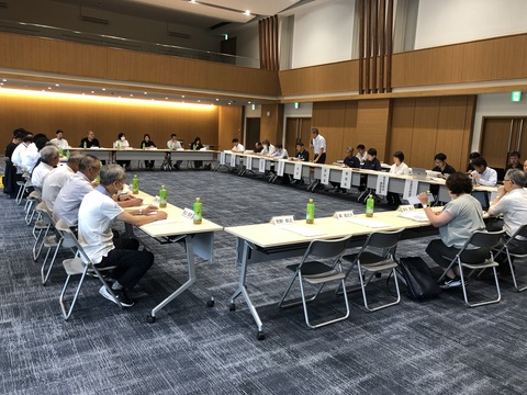 会場内全体を使用して長テーブルをロの字型に配置し、参加者が着席して資料を見ながら話し合っている様子の写真